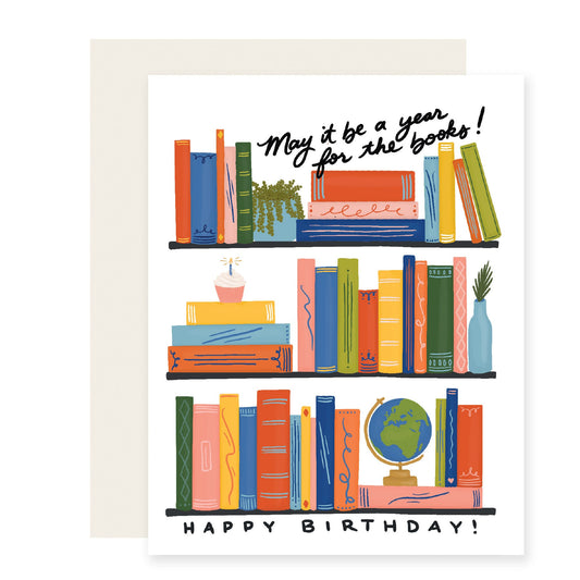 A delightful birthday card featuring beautifully illustrated bookshelves adorned with colorful books. Among the shelves, a special touch includes a birthday cake, a globe, and a house plant – a perfect celebration for the avid book lover. The card reads, 'May it be a year for the books. Happy Birthday! 