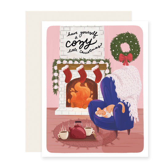 A beautifully illustrated Christmas card with a cozy scene: fireplace, snuggled-up cat, comfy chair, and hot beverages. Text reads 'Have yourself a cozy little Christmas'.