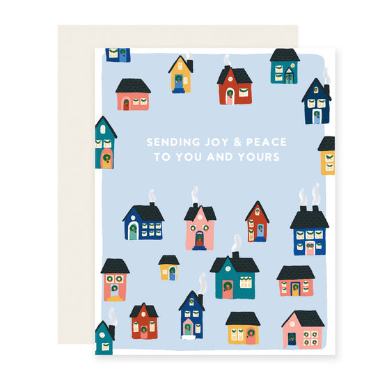A cozy holiday card with a light blue background. Illustrated with charming, colorful houses emitting smoke from their chimneys. The text reads 'Sending you joy and peace to you and yours'.